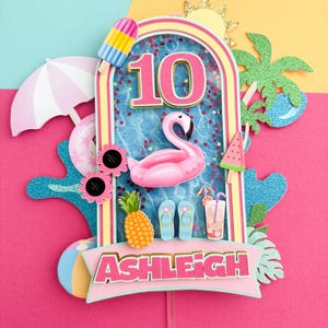 May include: A colorful birthday cake topper with a summer theme. The design includes a pink flamingo, a number 10, and the name "Ashleigh". Other elements include a popsicle, pineapple, and a cocktail.