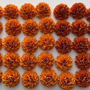 May include: An overhead shot displays a grid of orange paper flowers. Each flower has multiple layers of petals, creating a full, rounded shape. The flowers are a vibrant shade of orange, with slight variations in tone.