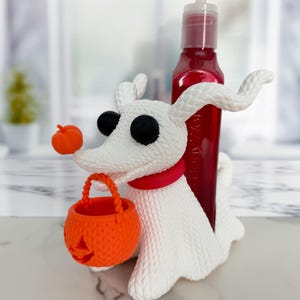 May include: A white ghost dog figure with a textured knit pattern, black eyes, and a red collar. The figure holds a small orange jack-o'-lantern bucket in its mouth and has a tiny orange pumpkin on its nose. A red soap dispenser is inserted behind.