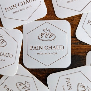 May include: White square stickers with rounded corners, featuring a brown hexagon design with a bread illustration and the words "PAIN CHAUD MADE WITH LOVE". The stickers are scattered on a wooden surface.