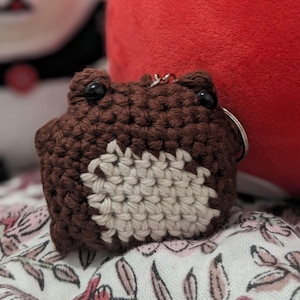 May include: A brown crocheted frog keychain with white crocheted details and black button eyes.