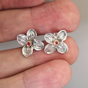 Op de afbeelding: Een paar zilveren oorbellen in de vorm van een bloem met een rode edelsteen in het midden van elke bloem.