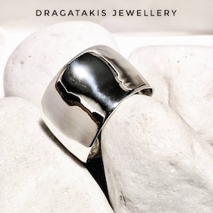 Può includere: Un anello largo in metallo argentato lucido con una parte superiore arrotondata e liscia. L'anello è su una superficie bianca e liscia.