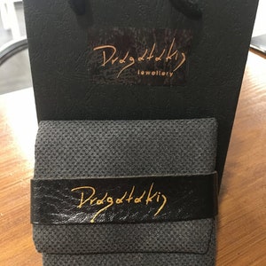 May include: A black gift bag with a gold logo that says "Dragottaki Jewellery". The bag is sitting on a wooden surface with a black fabric box that has a gold logo that says "Dragottaki" on the front.