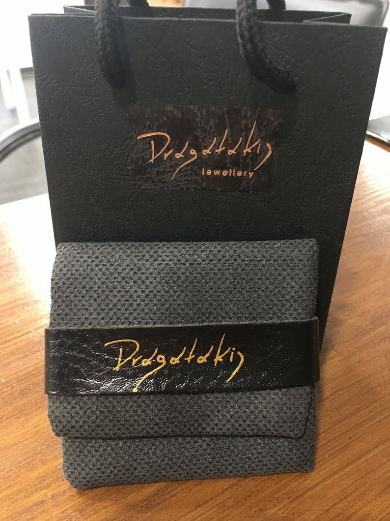 May include: A black gift bag with a gold logo that says "Dragottaki Jewellery". The bag is sitting on a wooden surface with a black fabric box that has a gold logo that says "Dragottaki" on the front.