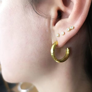 May include: Gold hoop earrings with a hammered texture. The earrings are worn in a person's ear with several small diamond studs.