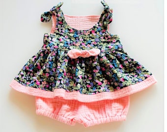 May include: A pink and black floral print baby romper with a bow. The romper has a ruffled skirt and a gathered bodice.