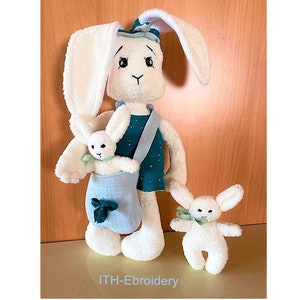 May include: Three white plush bunny toys. The largest bunny is wearing a teal and gold dress and carrying a smaller bunny in a light blue bag. The smallest bunny is standing on its own.