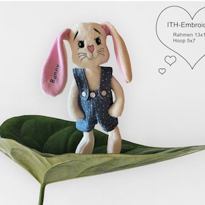 Puede incluir: Un conejito de peluche blanco que lleva un mono azul con lunares blancos. El conejito tiene orejas rosas con la palabra "Bunny" bordada en una oreja. El conejito está sentado en una hoja verde. La imagen incluye el texto "ITH-Embroidery Rahmen 13x18 Hoop 5x7".