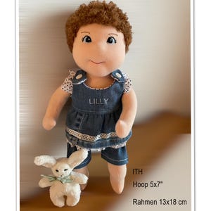 May include: A handmade doll with brown curly hair, wearing a denim outfit with the name "LILLY" embroidered on the front. A small white stuffed bunny with a green bow is next to the doll. The doll is approximately 30 cm tall.