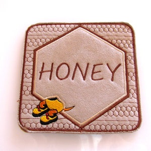 May include: A brown and beige embroidered coaster with a honeycomb pattern and the word "HONEY" in brown embroidery. A small embroidered bee with yellow and black accents is in the bottom right corner.