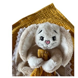 May include: A white plush bunny with embroidered eyes and a pink nose, holding a yellow and white polka dot blanket. The blanket has a white lace trim.