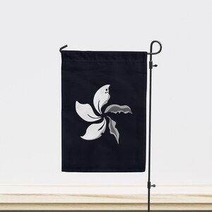 Puede incluir: Una bandera de jardín negra con un diseño de flor de Bauhinia blakeana blanca y gris. La bandera se muestra en un soporte de metal negro, contra una pared blanca y un fondo de hierba y una superficie de madera.