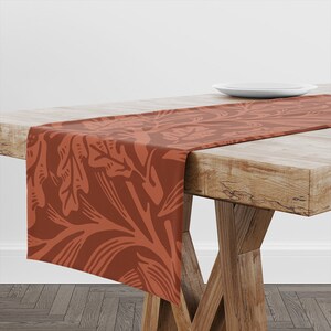 May include: A brown table runner with a floral pattern. The runner is made of a soft, woven fabric and features a repeating design of stylised leaves and stems.