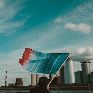 May include: A person holds a colorful striped flag in front of a large industrial complex with tall smokestacks and cylindrical tanks. The sky is blue with white clouds.