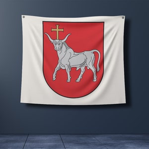 Puede incluir: Una bandera blanca con un escudo rojo que presenta un toro blanco con una cruz dorada en la cabeza. El toro está de pie sobre sus cuatro patas con la cabeza mirando hacia adelante. La bandera está colgada en una pared.
