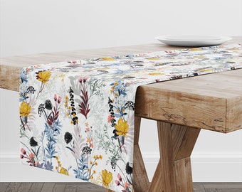 May include: A white table runner with a floral pattern featuring yellow, blue, red, and black flowers. The runner is on a wooden table.