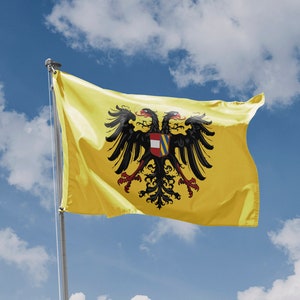 Op de afbeelding: Een gele vlag met een zwarte dubbelkoppige adelaar en een rood-wit schild in het midden. De vlag wappert in de wind tegen een blauwe lucht met witte wolken.