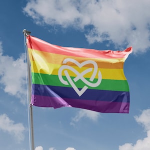 May include: A rainbow pride flag with a white heart and knot symbol in the centre, waving in the wind against a blue sky with white clouds.
