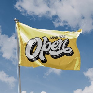 Puede incluir: Una bandera amarilla con el texto "We're Open" en letras blancas y negras. La bandera ondea al viento contra un cielo azul con nubes blancas.
