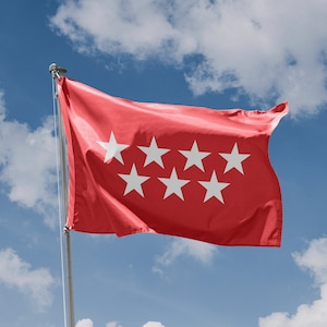 Op de afbeelding: Een rode vlag met zeven witte vijfpuntige sterren, wapperend in de wind tegen een blauwe lucht met witte wolken.
