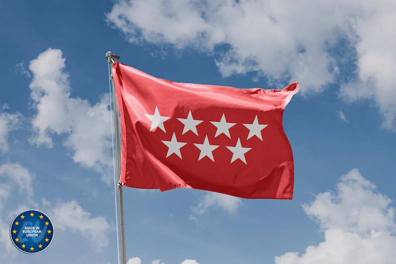 Op de afbeelding: Een rode vlag met zeven witte vijfpuntige sterren, wapperend in de wind tegen een blauwe lucht met witte wolken.