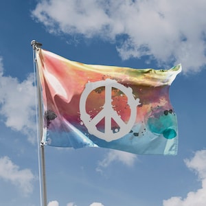 May include: A colorful flag with a peace symbol against a blue sky with clouds. The flag features a tie-dye design with a white peace sign in the center. The flag is waving in the wind.