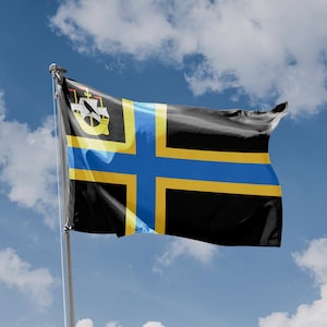 Puede incluir: Una bandera negra y amarilla con una cruz azul y un diseño de barco blanco en la esquina superior izquierda. La bandera ondea al viento contra un cielo azul con nubes blancas.