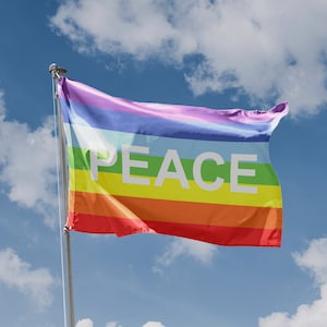 May include: A rainbow peace flag with the word "PEACE" in white letters on a white stripe, waving in the wind against a blue sky with white clouds.