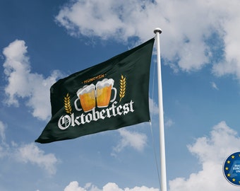 Op de afbeelding: Een groene vlag met de tekst "Oktoberfest" en twee bierglazen met de tekst "München" erop. De vlag wappert aan een witte mast tegen een blauwe lucht met witte wolken.
