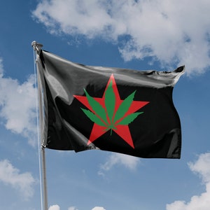 Puede incluir: Una bandera negra con una estrella roja de cinco puntas que presenta una hoja verde de marihuana en el centro. La bandera ondea al viento contra un cielo azul con nubes blancas.