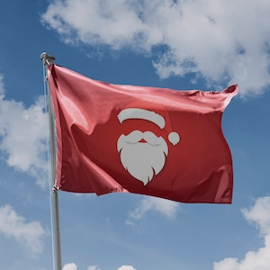 Peut inclure: Un drapeau rouge avec un visage de Père Noël blanc dessus, flottant au vent contre un ciel bleu avec des nuages blancs.