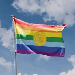 Könnte beinhalten: Eine Regenbogen-Pride-Flagge mit einem goldenen kreisförmigen Design in der Mitte, die an einem blauen Himmel mit weißen Wolken weht.