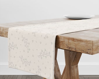May include: A beige table runner with a delicate floral pattern in gray. The runner is on a wooden table with a white plate on top.