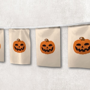 Könnte beinhalten: Eine Girlande aus fünf beigefarbenen Stofffahnen mit aufgedruckten orangefarbenen Kürbisgesichtern. Die Fahnen sind mit einer weißen Schnur zusammengebunden.