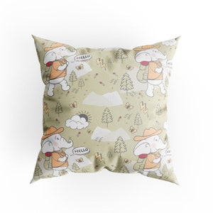 Puede incluir: Una almohada verde con un elefante blanco que lleva un sombrero y una mochila. El elefante camina por un bosque con árboles, nubes y montañas. El texto "Hello" está en un globo de diálogo sobre el elefante.