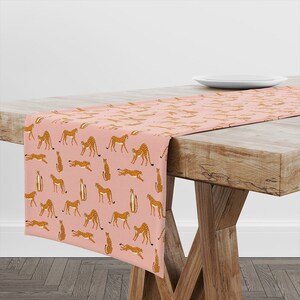 May include: A pink table runner with a repeating pattern of brown cheetahs in various poses. The runner is on a wooden table with a white plate on the table.