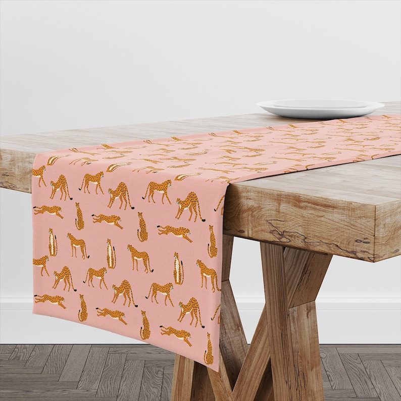 May include: A pink table runner with a repeating pattern of brown cheetahs in various poses. The runner is on a wooden table with a white plate on the table.