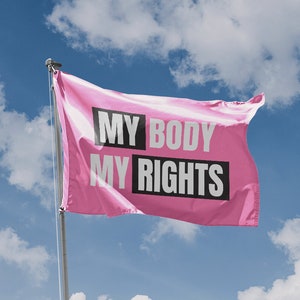 May include: A pink flag with the text "MY BODY MY RIGHTS" in white letters on a black background, waving in the wind against a blue sky with white clouds.