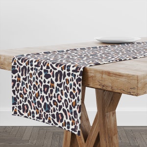 Puede incluir: Un camino de mesa blanco con un estampado de leopardo en tonos marrones, rosas y azules. El camino de mesa está sobre una mesa de madera.