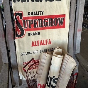 May include: A vintage white burlap sack with red and black text that reads "Reneau's Quality Supergrow Brand Alfalfa 50 lbs. Net 22.68 Kilos". The sack is sitting on a wooden chair with a red wire basket filled with more burlap sacks.
