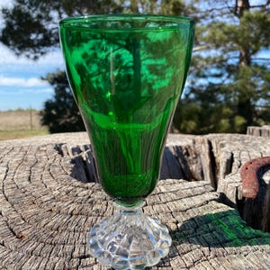 May include: A vibrant green glass goblet with a clear, textured base. The goblet's bowl is a deep emerald hue, reflecting the surrounding environment. The stem is clear, and the goblet sits on a weathered wooden surface, with a horseshoe in the background.