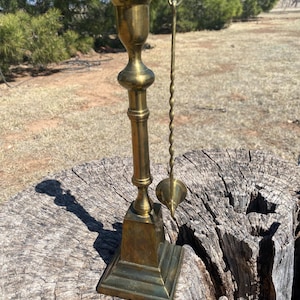 May include: A brass candlestick with a square base, a tall, slender stem, and a cup-shaped top. A matching brass candle snuffer hangs on the side. The candlestick is placed on a weathered tree stump.
