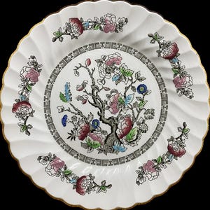 May include: A white ceramic plate with a scalloped edge and a gold-trimmed rim. The center features a detailed tree illustration with pink, blue, and green floral accents, surrounded by a black geometric border and floral sprigs on the rim.