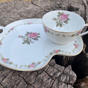 May include: White porcelain teacup and saucer set with a pink rose design. The cup and saucer have gold trim and a floral border. The saucer has a unique shape with a handle. A classic tea set.