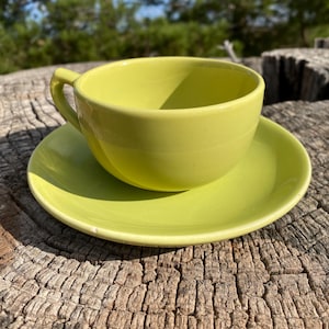 May include: A lime green teacup and saucer set. The cup has a curved handle and sits on a matching saucer. The set is made of ceramic and is placed on a weathered wooden surface, with a blurred natural background.