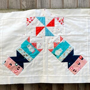 May include: A handmade quilt featuring a repeating pattern of colorful blocks. The blocks include images of polar bears, crowns, and geometric shapes in shades of blue, red, orange, and white. The quilt is set against a cream-colored background.