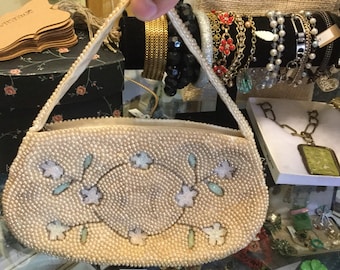 Könnte beinhalten: Eine weiße Perlenhandtasche mit Blumenmuster. Die Handtasche hat einen langen Riemen und besteht aus kleinen, weißen Perlen. Die Perlen sind in einem Muster aus Blumen und Blättern angeordnet.