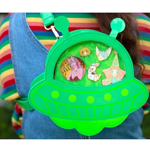 May include: A bright green, UFO-shaped crossbody bag with a clear window displaying several colorful pins. The bag has a strap and is worn over a denim overall. The pins include cartoon characters and the word "Hello".