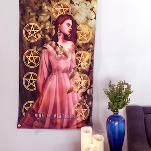 Puede incluir: Un tapiz de pared con la carta del tarot Nueve de Oros. La obra de arte representa a una mujer con un vestido rosa rodeada de pentáculos dorados y follaje. El texto "NINE OF PENTACLES" es visible en la parte inferior. El tapiz está colgado en una pared blanca.